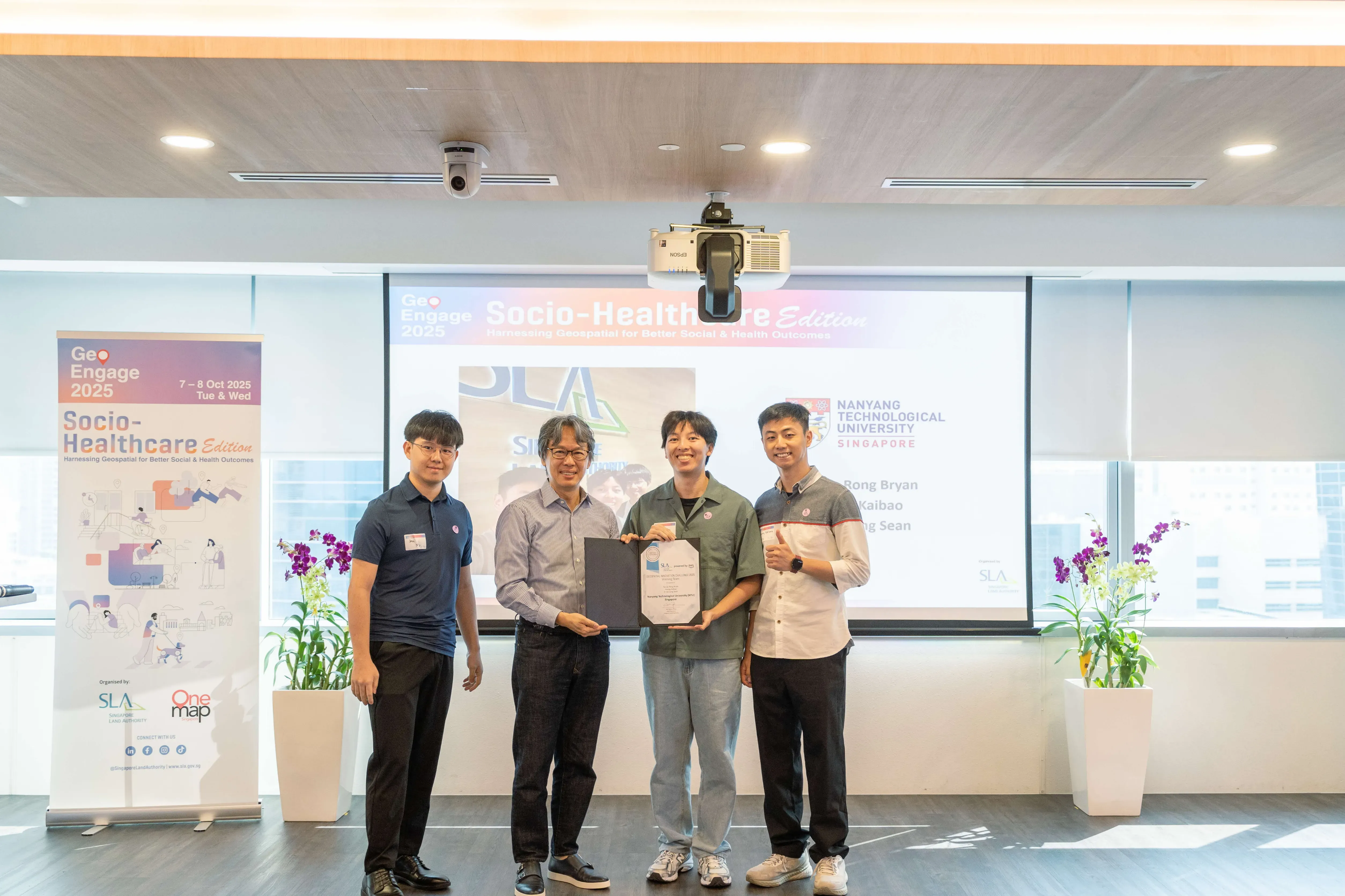 SLA CE Mr Calvin Phua with Team 1 at a GeoEngage 2025 Socio-Healthcare event, two holding an award certificate in front of a projector screen.